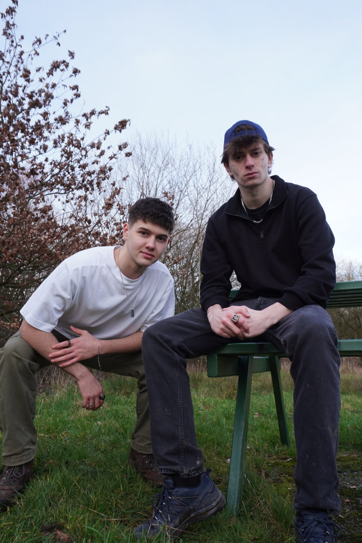 Two young men sitting on a green park bench outdoors under an overcast sky, with grass and trees in the background.