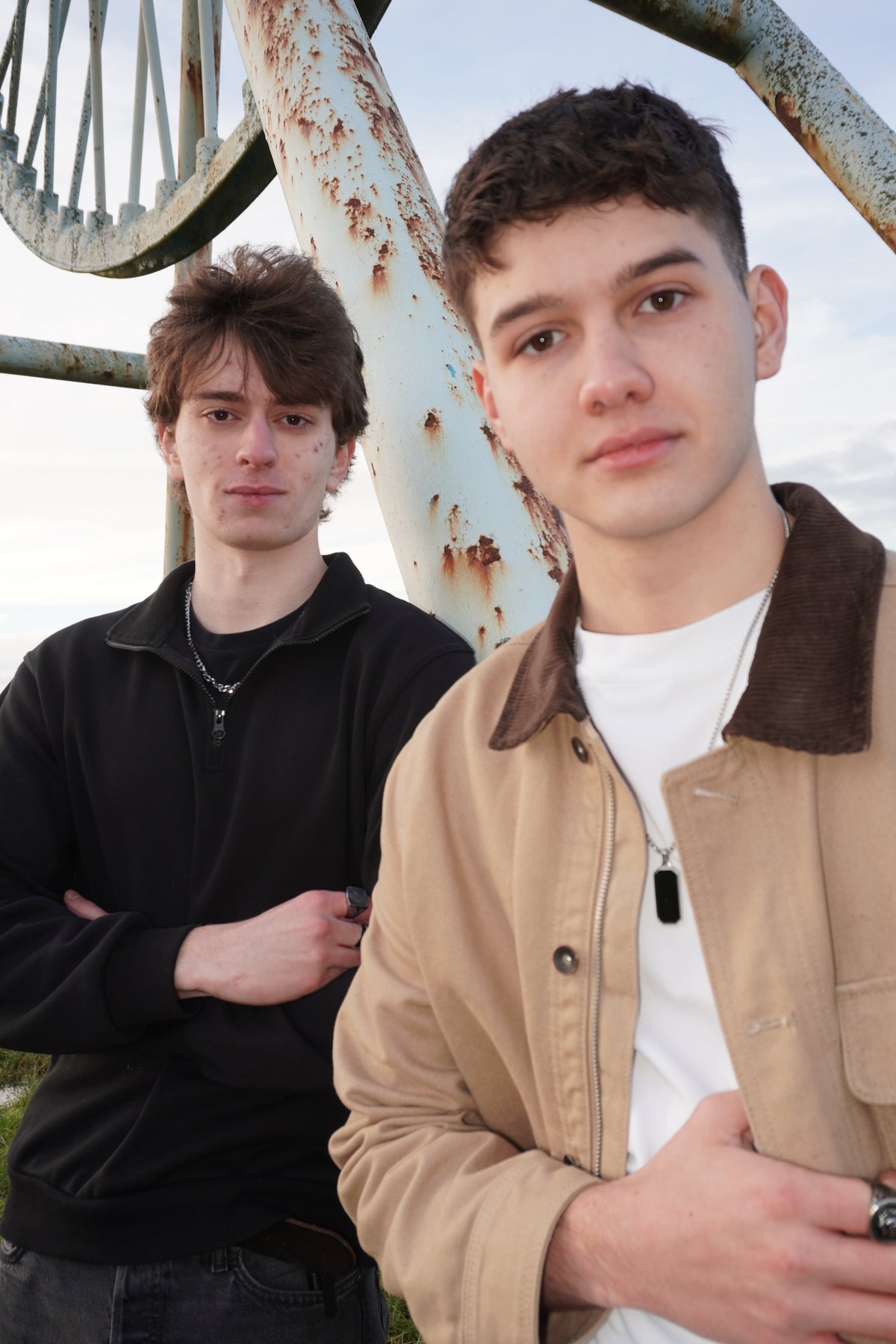 Two young men standing outdoors in front of a large rusted metal structure. The man in the foreground wears a beige jacket over a white t-shirt, and the man behind him wears a black zip-up hoodie.